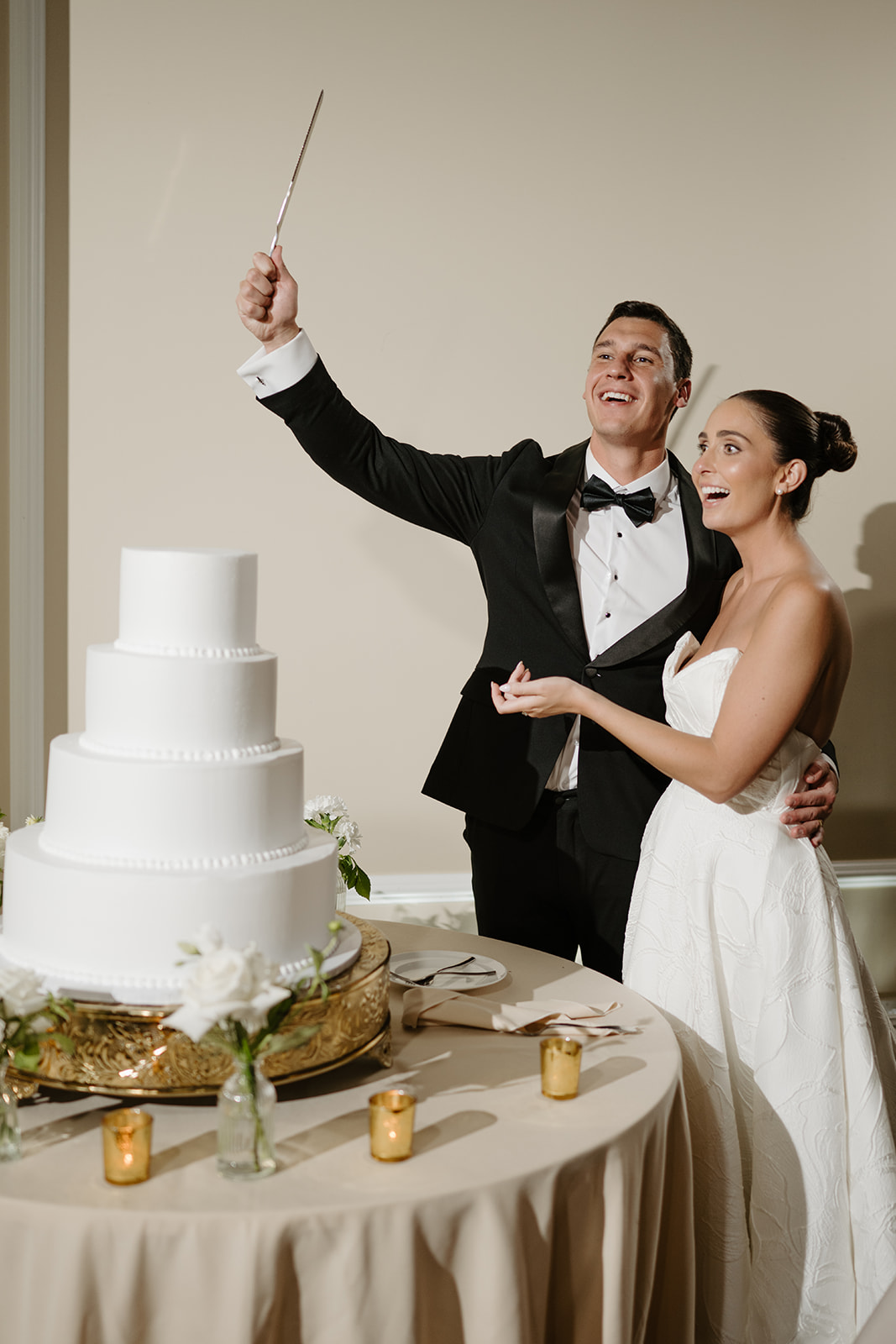 Newlyweds laughing as the groom holds the cake knife high in the air before cutting their four-tier cake.